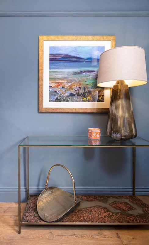 Blue-gray wall with framed landscape artwork, glass console table with lamp, decorative basket and candle