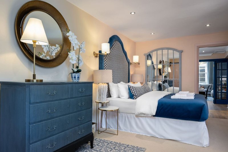 Spacious bedroom with blue upholstered headboard, white bedding, blue dresser, round mirror and ensuite bathroom visible
