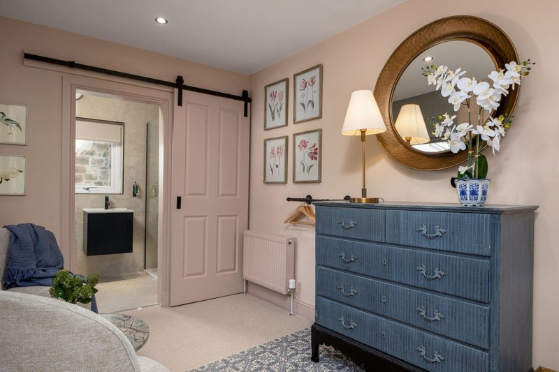 Bedroom with blue dresser, round mirror, white flowers, and ensuite bathroom visible through doorway