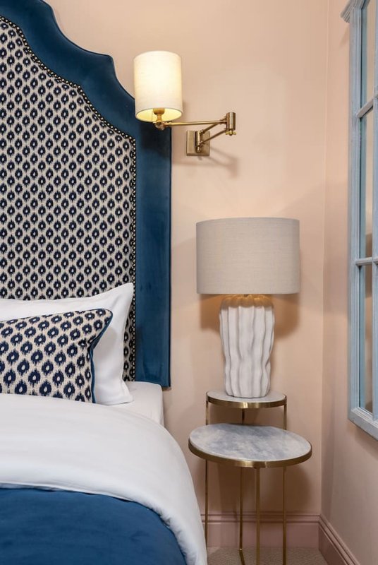 Bedroom corner with blue upholstered headboard, white bedding, wall sconce, and side table lamp