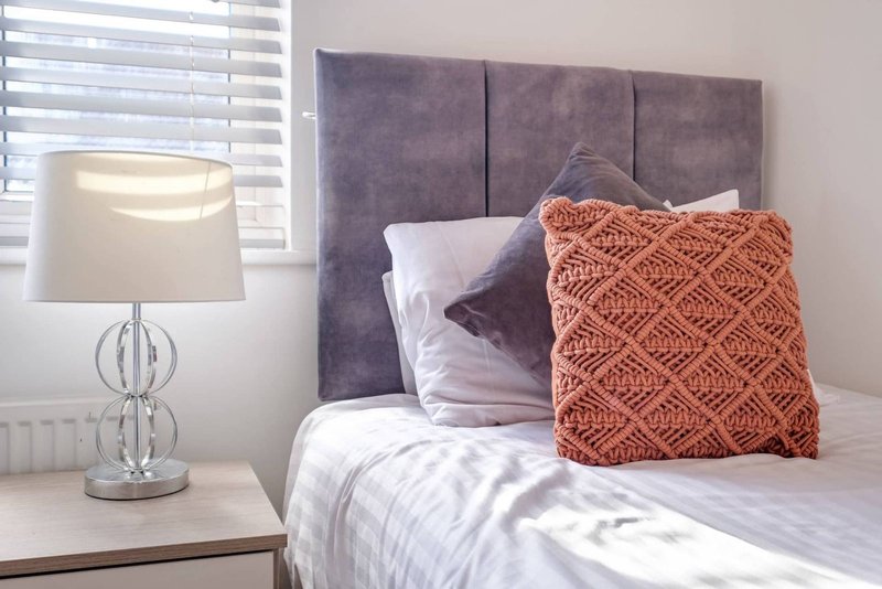 Bedroom with double bed, grey upholstered headboard, orange decorative pillow, bedside lamp and window