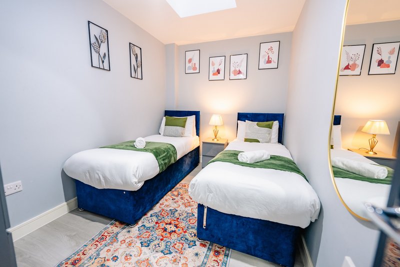 Bright twin bedroom with two blue beds, white pillows, green throws, bedside lamps and framed wall art