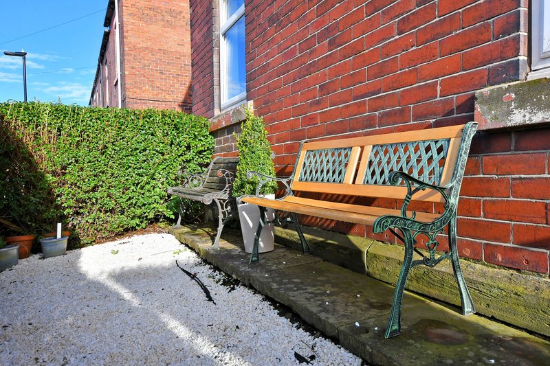 Brick building exterior with wooden bench, green hedge, potted plants and paved courtyard area