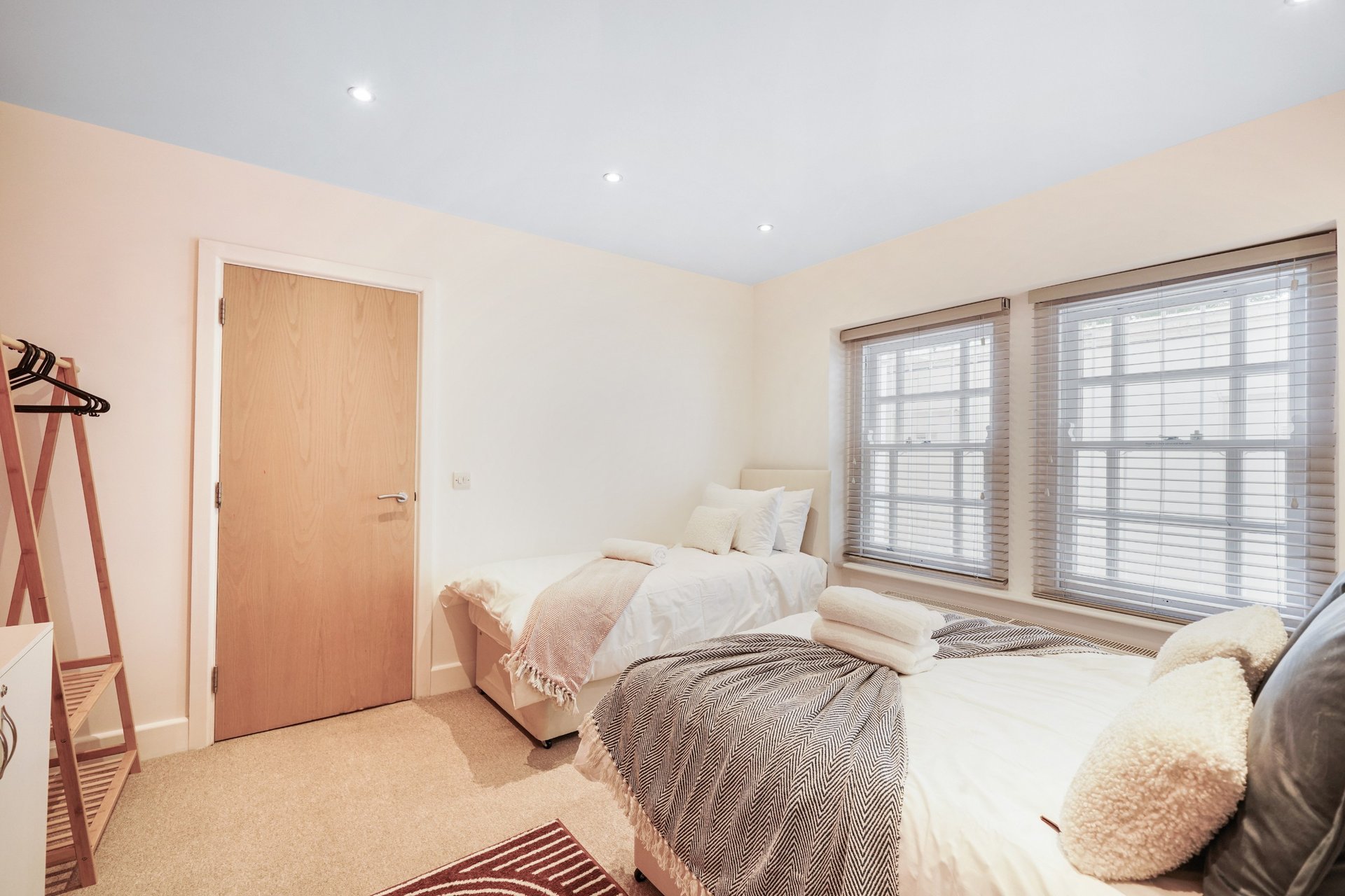 Bright bedroom with two single beds, wooden door, large windows with shutters, and neutral décor