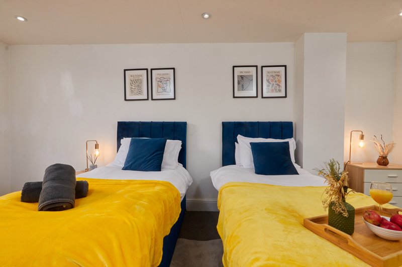 Twin bedroom with yellow bedding, blue headboards, white pillows, framed wall art, and bedside lamps