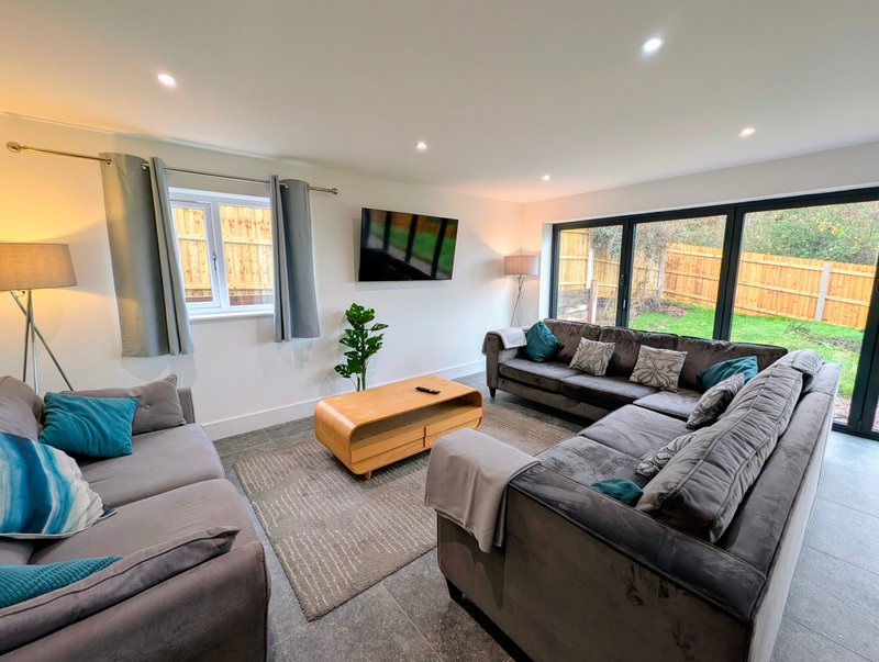 A spacious, modern living room with comfortable seating for larger groups, a wall-mounted Smart TV and a large coffee table. Full-height patio doors overlook the private garden.