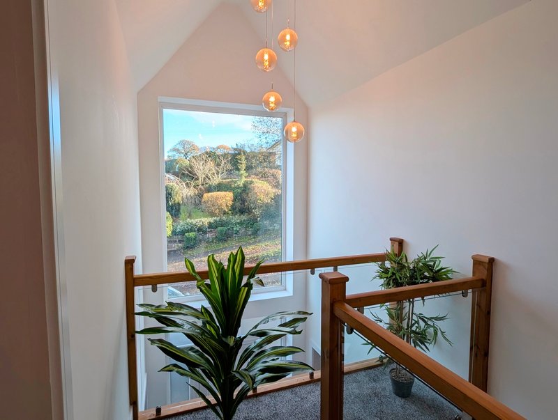 A bright and welcoming upstairs landing with a large feature window overlooking the garden, complemented by wooden and glass balustrades, greenery and stylish hanging lights.
