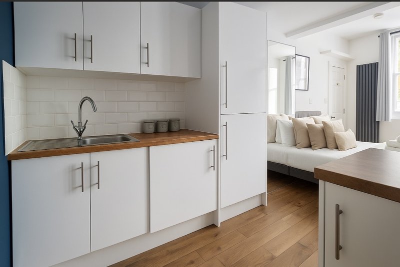 Modern kitchenette with white cabinetry, wooden countertop, and view into bedroom with bed and pillows
