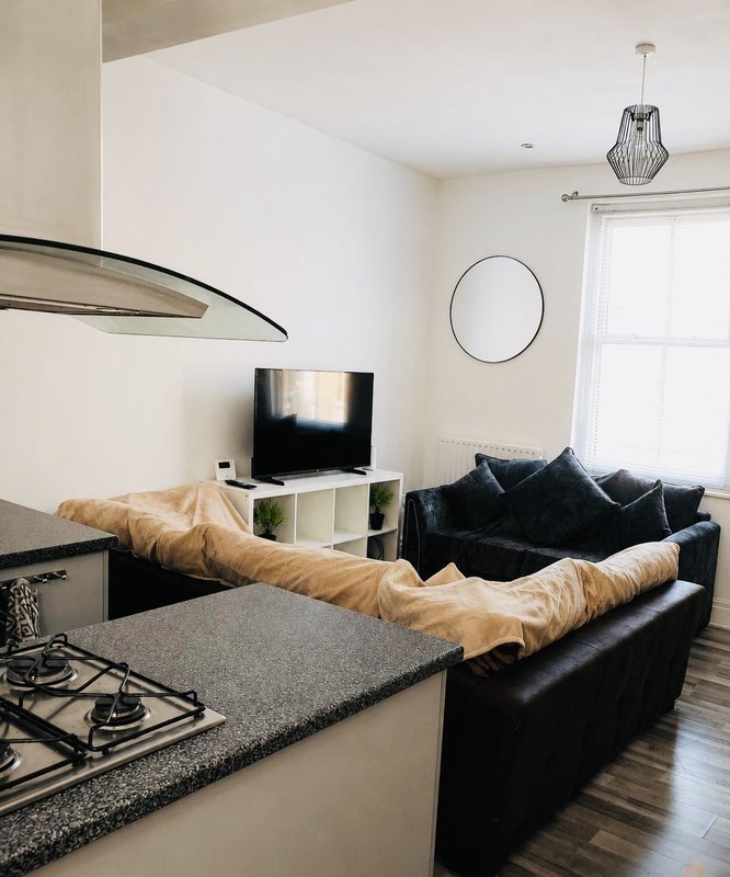 Open-plan living room with black sofa, TV on stand, kitchen counter with gas hob, and pendant light