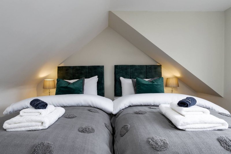 Twin beds with green velvet headboards, white pillows, gray bedding and bedside lamps in attic bedroom