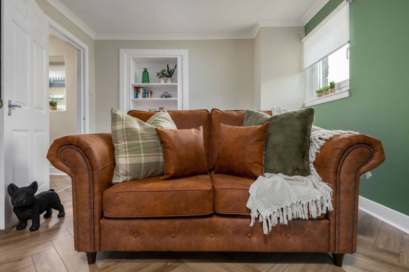 Living room with brown leather Chesterfield sofa, decorative cushions, throw blanket, and small black dog on wooden floor