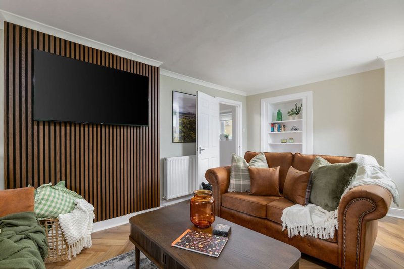 Living room with brown leather sofa, wooden coffee table, TV on wall, and large windows with wooden blinds