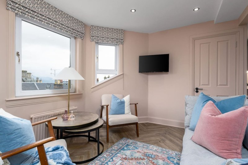 Bright bedroom with double bed, blue and pink cushions, wooden chair, side table and wall-mounted TV