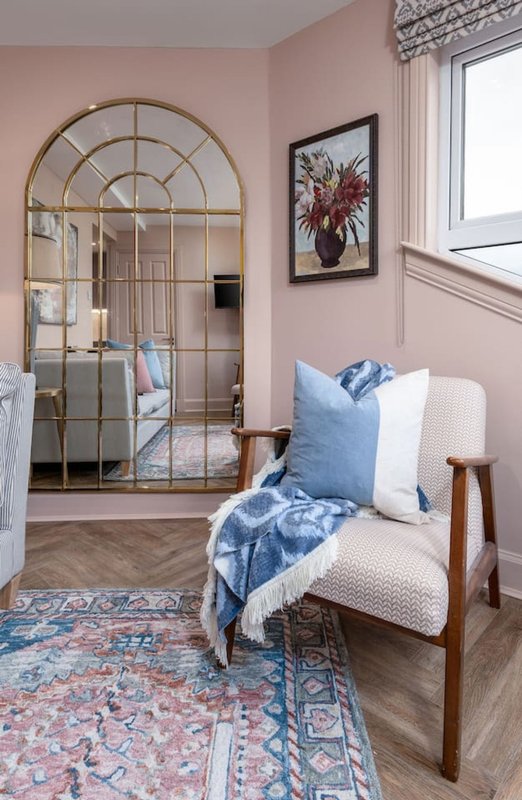 Bedroom with wooden chair, blue cushions, arched mirror, and framed floral artwork on pink wall