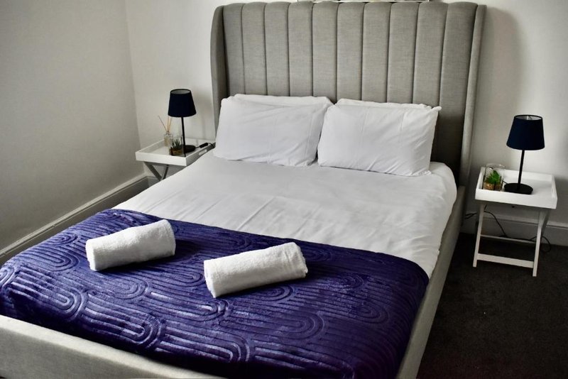Modern bedroom with grey upholstered headboard, white pillows, purple blanket and rolled towels