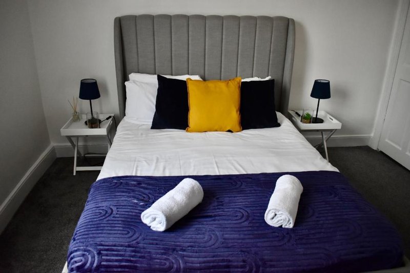 Modern bedroom with grey upholstered headboard, white bedding, yellow and black cushions, rolled towels, and bedside lamps