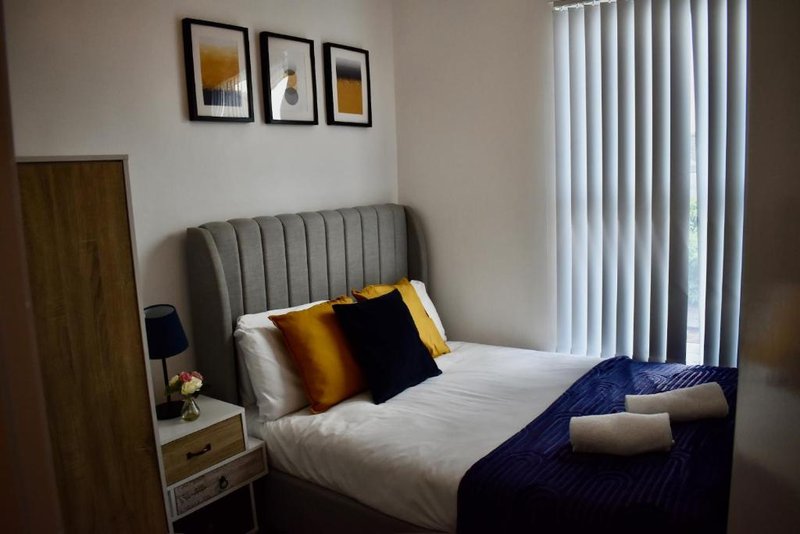 Modern bedroom with grey upholstered bed, white bedding, colourful cushions, and vertical blinds