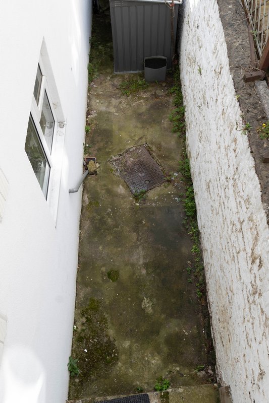 Narrow side passage between white-rendered and brick walls with moss-covered concrete ground