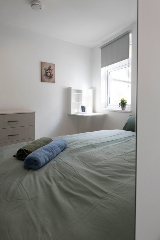 Bright bedroom with double bed, grey bedding, wooden dresser and window with blind