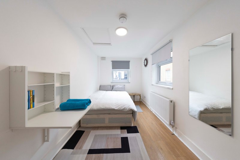 Narrow modern bedroom with single bed, desk, shelving, large mirror and wooden flooring