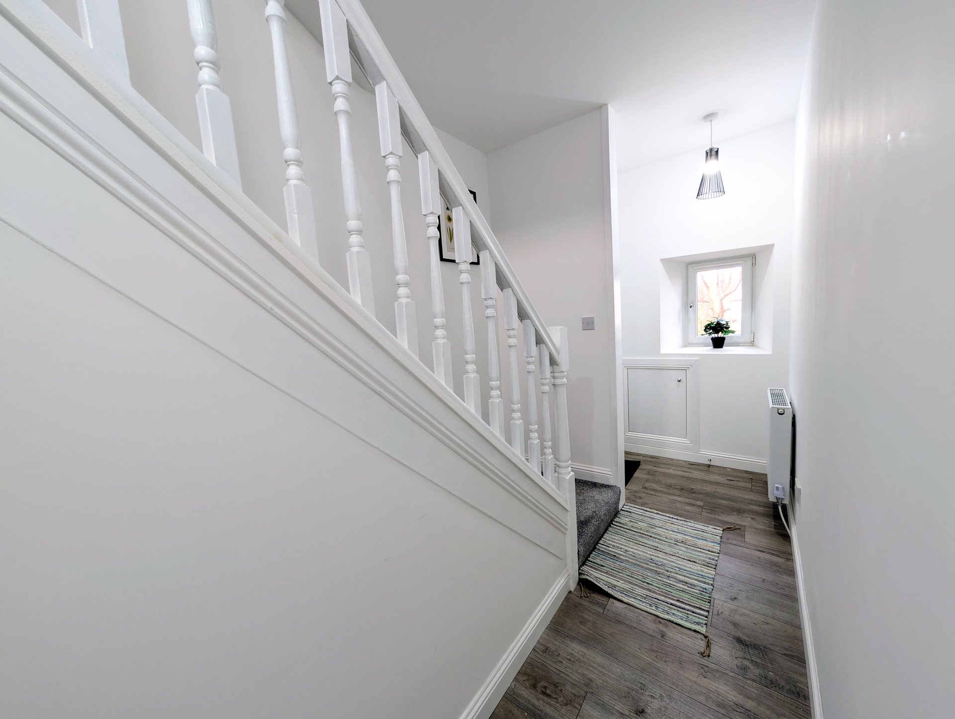 Bright hallway with white staircase, wooden flooring, and small window at end