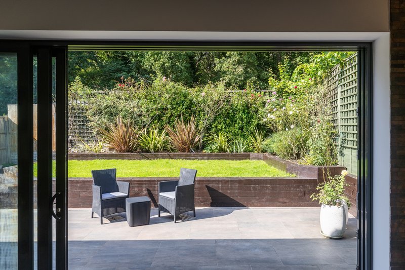 View through glass doors to patio with seating, raised garden bed and landscaped grounds