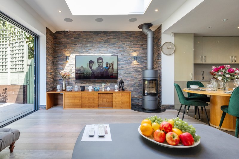 Open-plan living space with brick feature wall, wood-burning stove, dining table with fresh fruit, and kitchen area