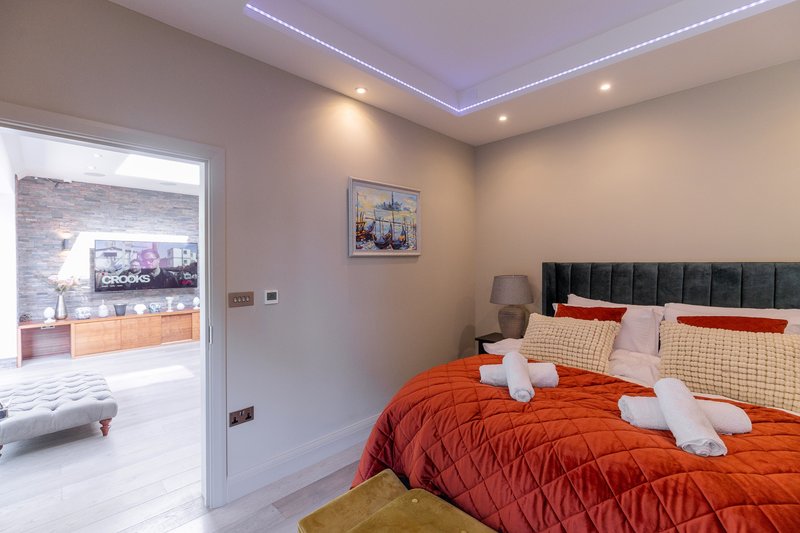 Modern bedroom with orange bedding, dark headboard, LED ceiling lights and open doorway to adjacent room