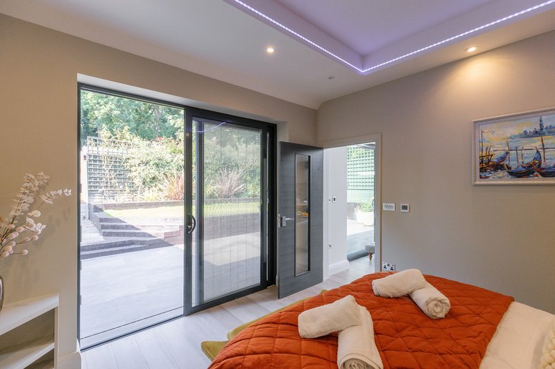 Modern bedroom with orange bedding, white towels, sliding glass doors to garden, and recessed ceiling lights