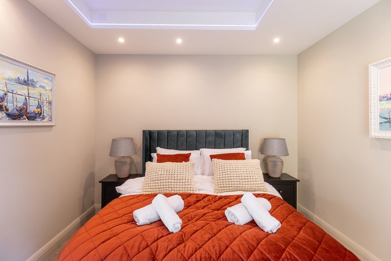 Modern bedroom with double bed, orange bedding, white towels, grey upholstered headboard and bedside lamps