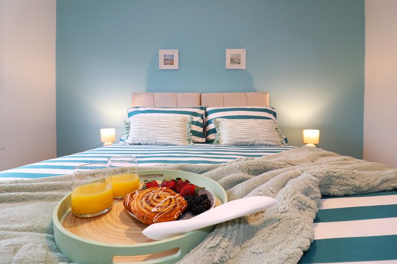 Bedroom with double bed, blue headboard, striped pillows, bedside lamps, and breakfast tray with juice and pastries