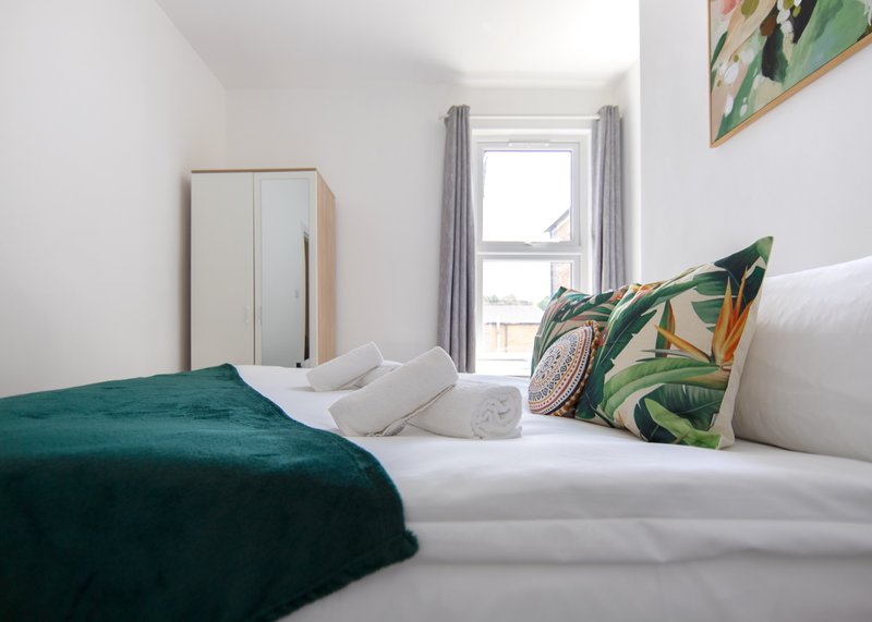 Bedroom with double bed, green bedding, patterned cushions, wooden wardrobe and window with grey curtains