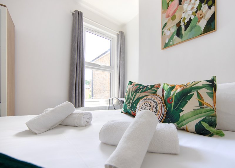 Bright bedroom with double bed, rolled white towels, patterned cushions and window with grey curtains