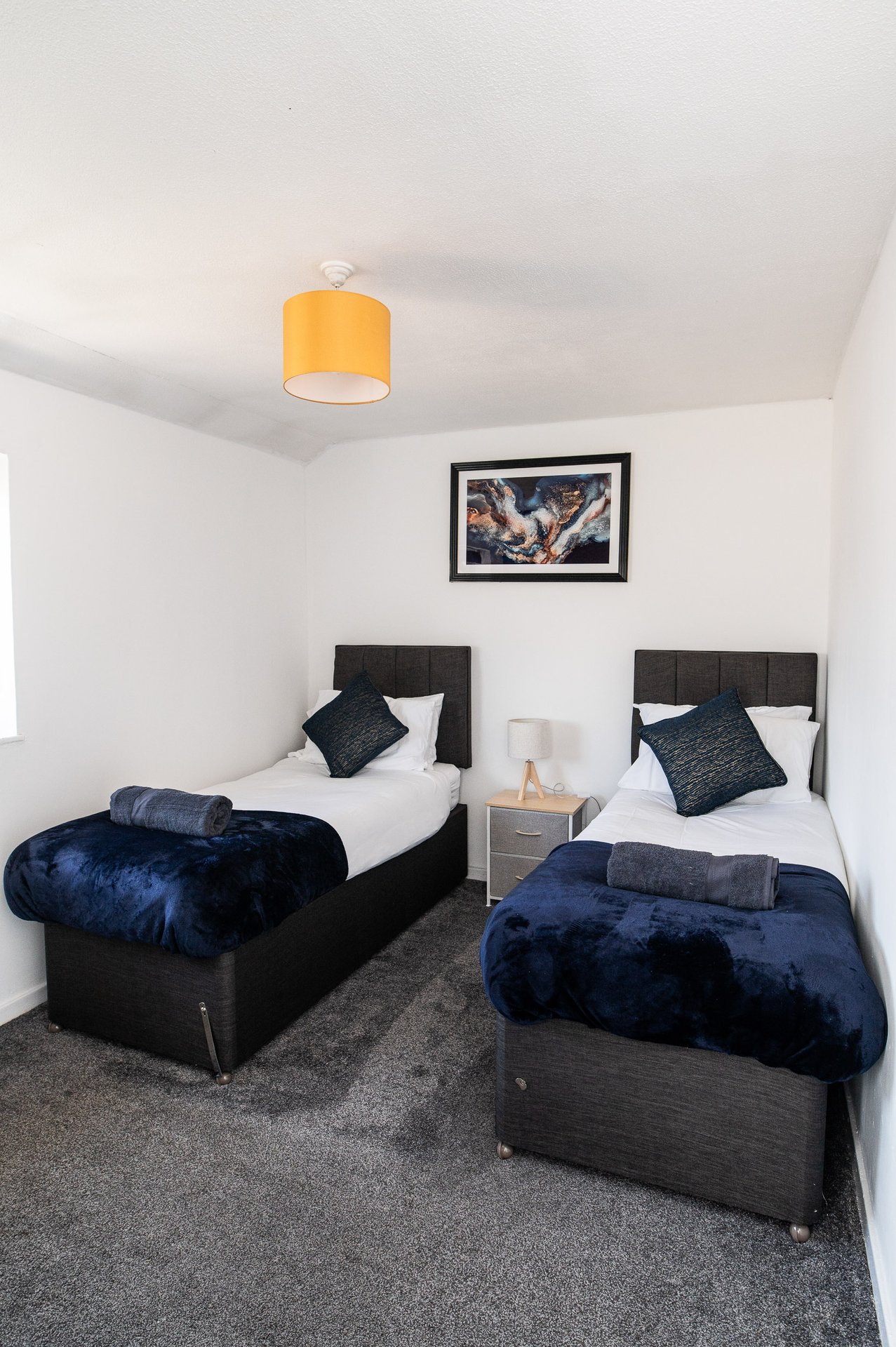Twin bedroom with black beds, blue velvet throws, white pillows, framed artwork and orange ceiling light