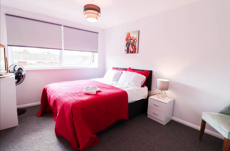 Bright bedroom with single bed, red bedding, pink blinds, bedside lamp and nightstand