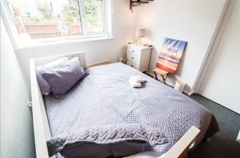 Small bedroom with single bed, purple pillow, white bedside table, and window with natural light