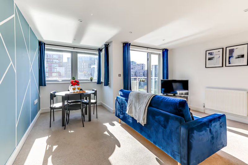 Modern open-plan living room with blue sofa, dining table, large windows and city views