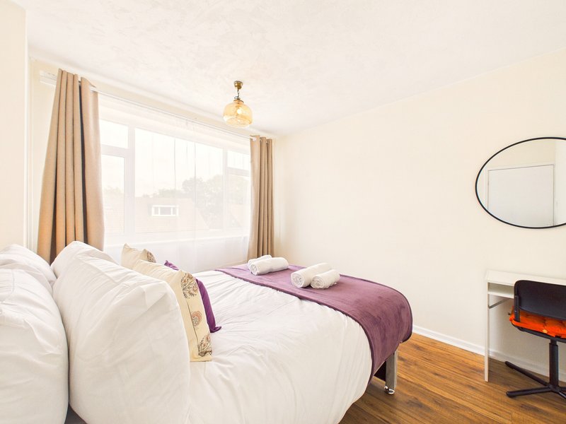 Bright bedroom with double bed, purple throw, large window with beige curtains and wooden flooring