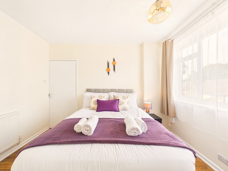 Bright bedroom with purple bedding, rolled towels, and large window with curtains