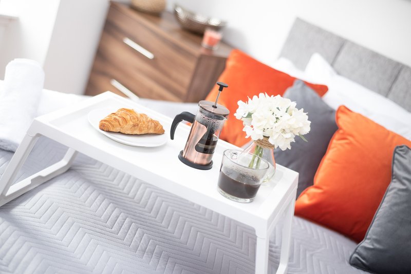 Bedroom with bed, white tray holding croissant, coffee pot, flowers and cushions