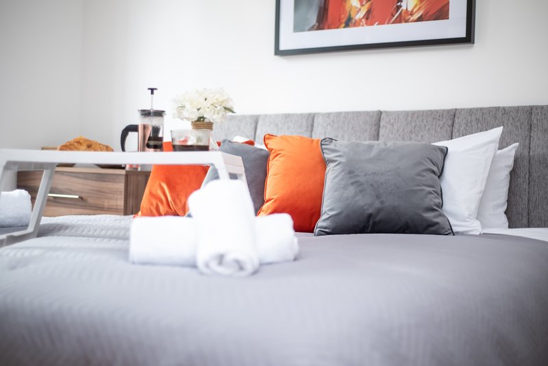 Modern bedroom with gray upholstered headboard, orange and gray cushions, white bedding, and framed wall art