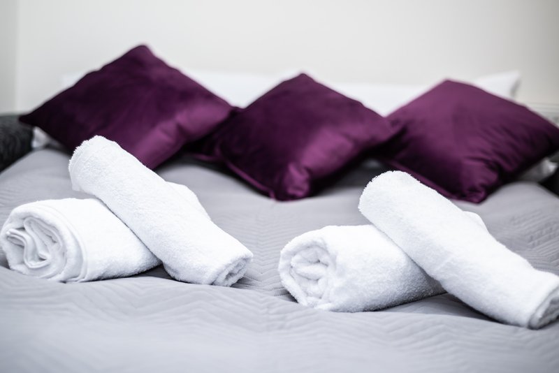 Bedroom bed with rolled white towels, purple and grey cushions arranged on headboard