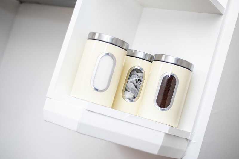 Three cream-colored storage canisters with metal lids on white shelf