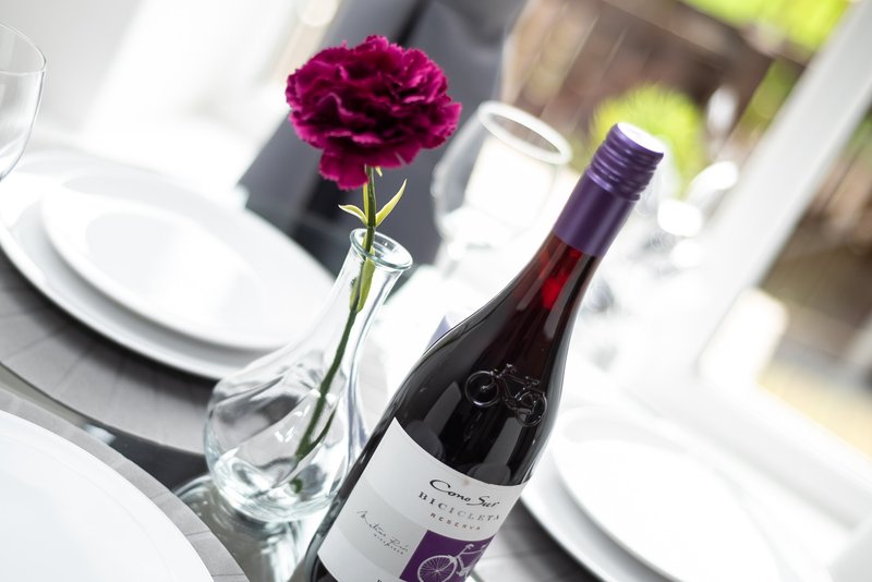 Dining table setting with wine bottle, glass vase with pink flower, and place settings