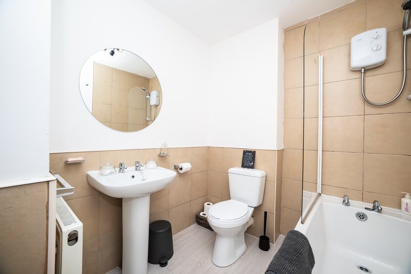 Bathroom with pedestal sink, toilet, bathtub, round mirror and beige tile walls