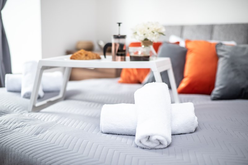 Bedroom with rolled white towels on bed, white tray table, orange and gray cushions, flowers