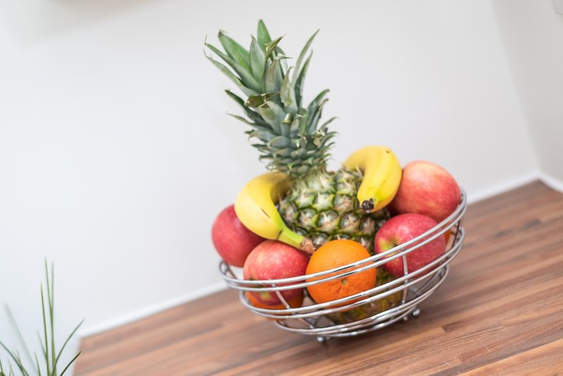 Wire fruit bowl with pineapple, bananas, apples and oranges on wooden table