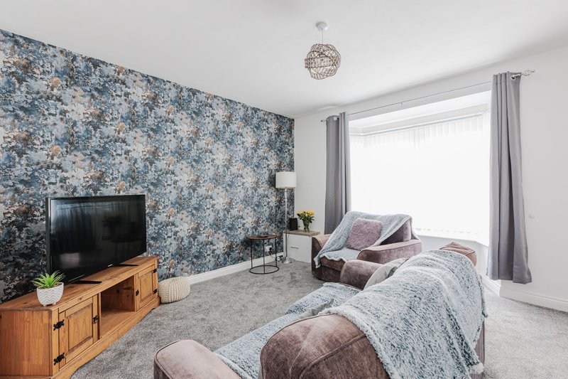 Bedroom with patterned blue wallpaper, wooden TV stand, bed with grey bedding and pink cushions, window with grey curtains