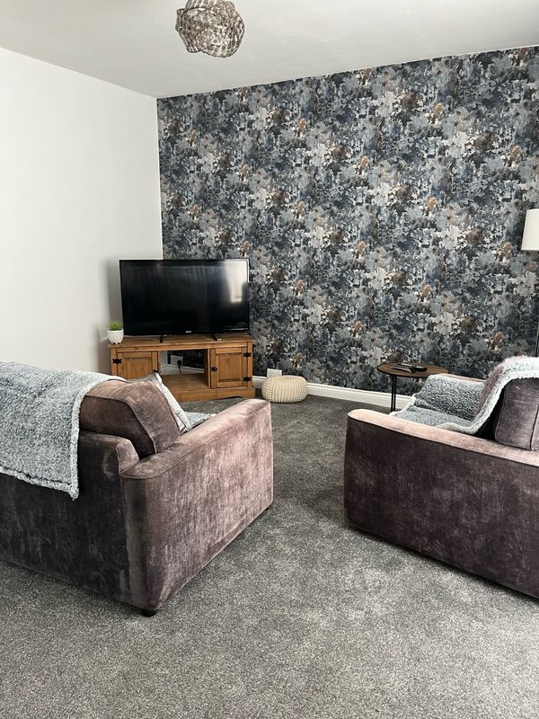 Living room with two upholstered armchairs, TV on wooden stand, floral wallpaper accent wall, and ceiling pendant light