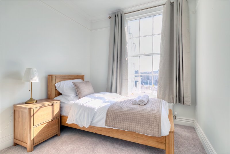 Bright bedroom with wooden bed frame, bedside table, lamp, and large window with grey curtains
